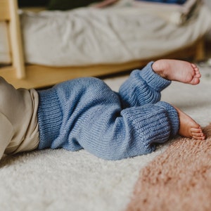 May include: A baby wearing a light blue knitted pants on a white and brown rug.