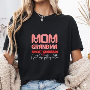 May include: Black t-shirt with pink text that reads "MOM GRANDMA GREAT GRANDMA" with heart accents. Below the text is the phrase "I just keep getting better" in a script font.