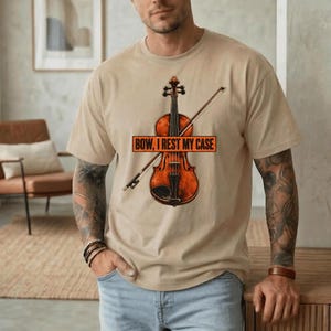 Könnte beinhalten: Ein beiges Kurzarm-T-Shirt mit einer Grafik einer braunen Holzgeige und einem Bogen. Über der Mitte der Geige befindet sich ein rechteckiges orangefarbenes Banner mit dem schwarzen Text "BOW, I REST MY CASE" in fetter Schrift.