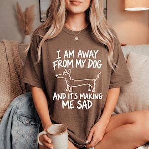 Peut inclure: Un t-shirt marron à manches courtes avec un dessin au trait blanc représentant un teckel et le texte "I AM AWAY FROM MY DOG AND IT'S MAKING ME SAD" imprimé en lettres blanches grasses et sans empattement au centre de la poitrine.