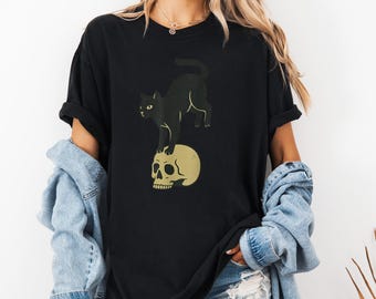 May include: Black t-shirt featuring a black cat standing on a golden skull. The graphic design is centered on the front of the shirt. The shirt has short sleeves and a relaxed fit, suitable for casual wear.