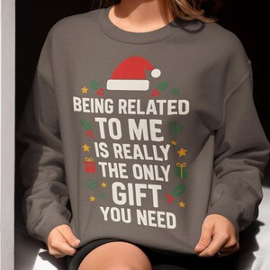 Peut inclure: Un sweat-shirt gris avec le texte "ÊTRE LIÉ À MOI EST VRAIMENT LE SEUL CADEAU DONT VOUS AVEZ BESOIN" en blanc. Le motif comprend un chapeau de Père Noël rouge, des étoiles, du houx et des cadeaux.