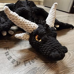 Może przedstawiać: Duża, ręcznie robiona czarna pluszowa zabawka w kształcie smoka z szarymi rogami i złotymi oczami. Smok leży na drewnianej podłodze, prezentując szczegółową fakturę i wzór szydełkowy. Pluszak to unikalny, ręcznie robiony przedmiot.