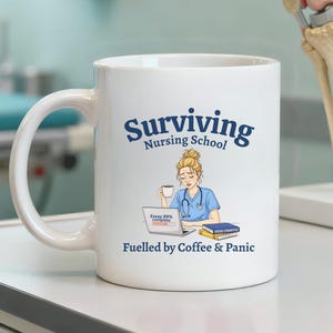 Peut inclure: Mug en céramique blanche avec l'inscription "Surviving Nursing School" en bleu marine. La tasse présente une illustration d'une infirmière avec un ordinateur portable et du café, avec le texte "Fuelled by Coffee & Panic".