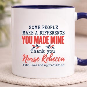 Può includere: Una tazza in ceramica bianca con interno e manico blu scuro. La parte anteriore presenta il testo: SOME PEOPLE MAKE A DIFFERENCE YOU MADE MINE Thank you Nurse Rebecca With love and appreciation, accentuato da un piccolo motivo grafico a foglie rosse e blu.