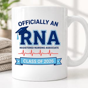 May include: A white ceramic mug featuring the blue text "OFFICIALLY AN RNA REGISTERED NURSING ASSOCIATE" with a blue graduation cap icon, a red EKG heartbeat line, and a light blue banner containing the white text "CLASS OF 2026".