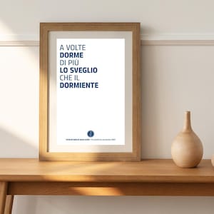 Peut inclure: Une impression encadrée avec un fond blanc et du texte bleu qui dit "A volte dorme di più lo sveglio che il dormiente". Le texte est dans une police moderne et est centré sur la page. Le cadre est en bois brun clair.