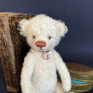 Può includere: Un orsacchiotto di peluche bianco con un naso rosa e un ciondolo floreale su una catenina d'argento. L'orsacchiotto è seduto su una superficie scura.