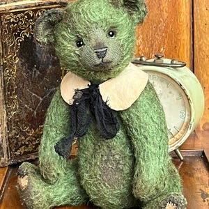 May include: A vintage-style green teddy bear with a distressed appearance. The bear has a cream-colored collar and a black bow. Its features include small black eyes and a stitched nose. The bear is sitting on a wooden surface, with a vintage clock and book in the background.