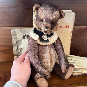 Puede incluir: Un oso de peluche de estilo vintage con un aspecto desgastado. El oso es principalmente marrón con un cuello blanco y negro. Está sentado frente a libros antiguos y una fotografía vintage.
