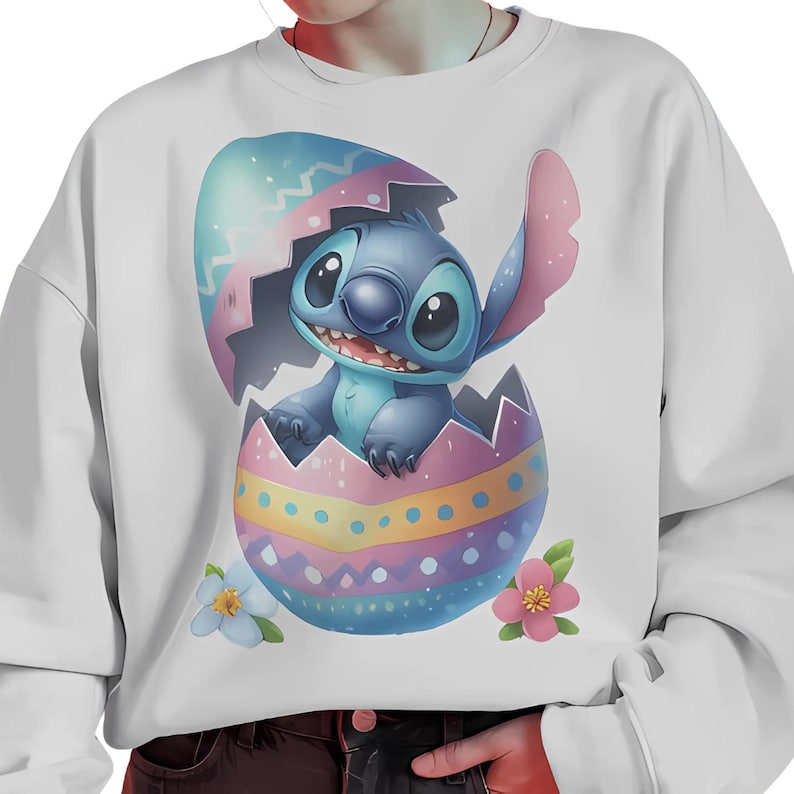 May include: A white sweatshirt with a blue and purple cartoon character in an Easter egg. The character has large eyes and a big smile. The egg is decorated with yellow, pink, and blue stripes and dots.