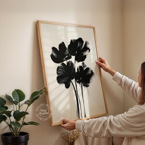 May include: A framed art print featuring a black floral design on a cream background. The artwork showcases bold brushstrokes, creating a modern and abstract aesthetic. The frame is made of light-colored wood, adding a natural touch to the piece.
