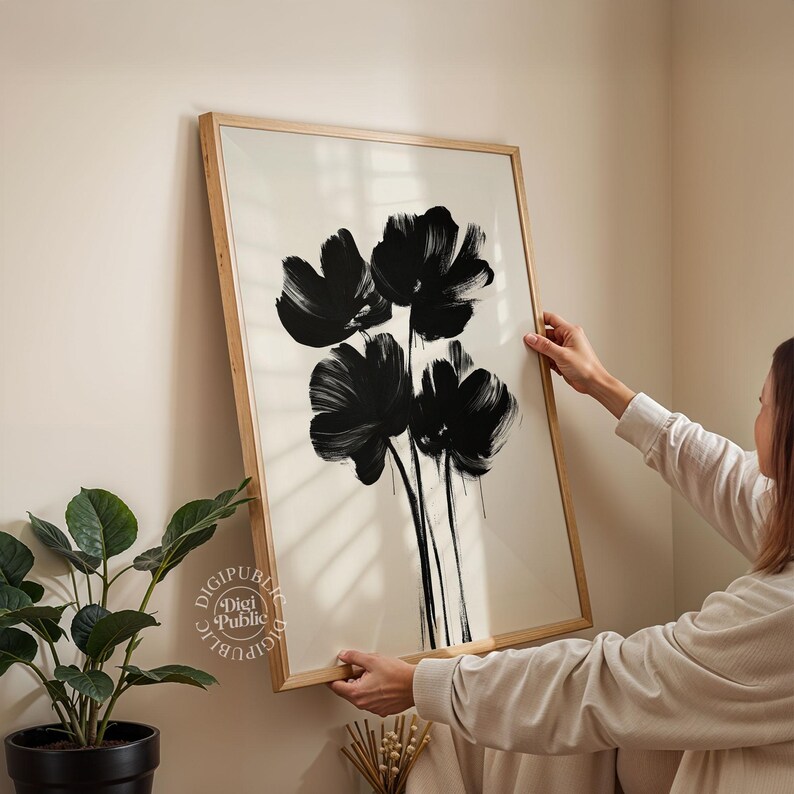 May include: A framed art print featuring a black floral design on a cream background. The artwork showcases bold brushstrokes, creating a modern and abstract aesthetic. The frame is made of light-colored wood, adding a natural touch to the piece.