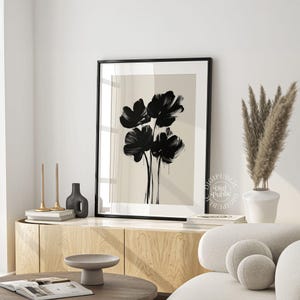 May include: Black and white floral art print in a black frame. The artwork features a minimalist design of five black flowers against a beige background. The print is displayed on a wooden cabinet, with decorative items and a sofa in the background.