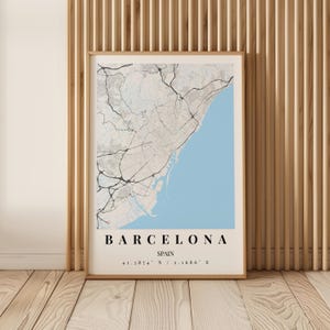 Pode incluir: Uma impressão de arte de parede emoldurada com um mapa de Barcelona, Espanha. O mapa exibe redes rodoviárias pretas sobre um fundo terrestre cinza claro, com o Mar Mediterrâneo em azul claro. O texto diz BARCELONA, SPAIN, 41.3874° N / 2.1686° E.