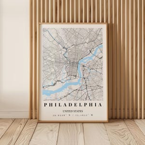 May include: A framed wall art print featuring a detailed map of Philadelphia, United States. The map displays grey street grids and a light blue river, with the text PHILADELPHIA, UNITED STATES, and coordinates 39.9526 N / 75.1652 W printed below.