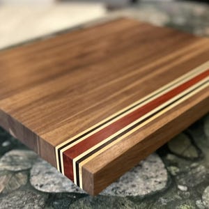 May include: A rectangular wooden chopping board crafted from dark brown walnut wood. The board features a decorative inlay stripe running across the surface, composed of a wide reddish-brown band flanked by thin black and cream-coloured accent strips.