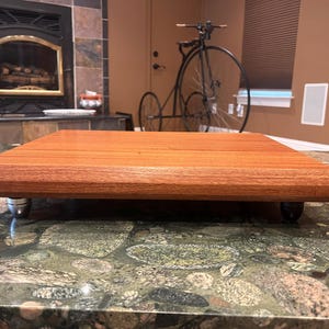 May include: A rectangular wooden platform with a smooth, reddish-brown grain finish and beveled edges. The platform is elevated by four small, cylindrical feet, with two silver-toned feet visible at the front corners and two dark feet at the back.