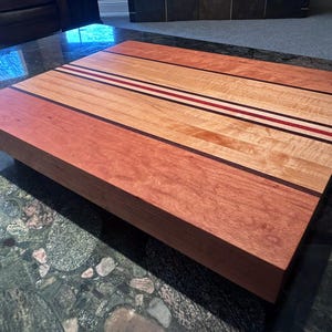 May include: A rectangular wooden coffee table with a multi-toned design. The table features a reddish-brown border, a light-colored central section, and thin stripes of red, white, and dark brown. The table is made of wood.