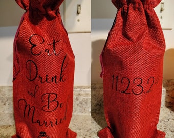 May include: Red burlap wine bottle bag with black text that reads "Eat Drink and Be Married" and two rings. The other side of the bag has the date "11.23.22" written in black.