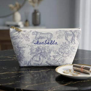 May include: A white and blue toiletry bag with a gold zipper. The bag features a tiger design and the name "Isabella" in blue script. The bag is on a black surface next to a small plate with jewelry and lipstick.
