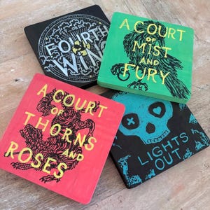 Puede incluir: Cuatro posavasos cuadrados con diseños temáticos de libros. Un posavasos negro presenta el texto "FOURTH WING". Un posavasos verde muestra "A COURT OF MIST AND FURY". Un posavasos rojo dice "A COURT OF THORNS AND ROSES". Un posavasos negro tiene una calavera y las palabras "LIGHTS OUT".