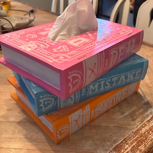 May include: Three brightly coloured tissue box covers, each designed to look like a book. The top one is pink and says "THE DEAL", the middle one is blue with "THE MISTAKE", and the bottom one is orange with "THE SCORE". White tissues protrude.
