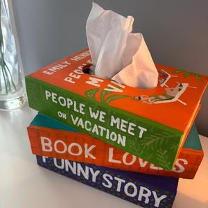Puede incluir: Tres cubiertas de caja de pañuelos pintadas a mano, diseñadas para parecerse a libros. La caja superior es naranja con el texto "People We Meet on Vacation". La caja del medio es turquesa y naranja con el texto "Book Lovers". La caja inferior es morada con el texto "Funny Story".
