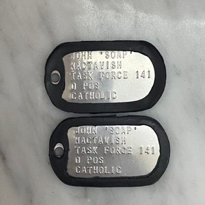 May include: Two silver dog tags with black rubber edging. The text on the tags reads "JOHN SOAP MACTAVISH TASK FORCE 141 O POS CATHOLIC".