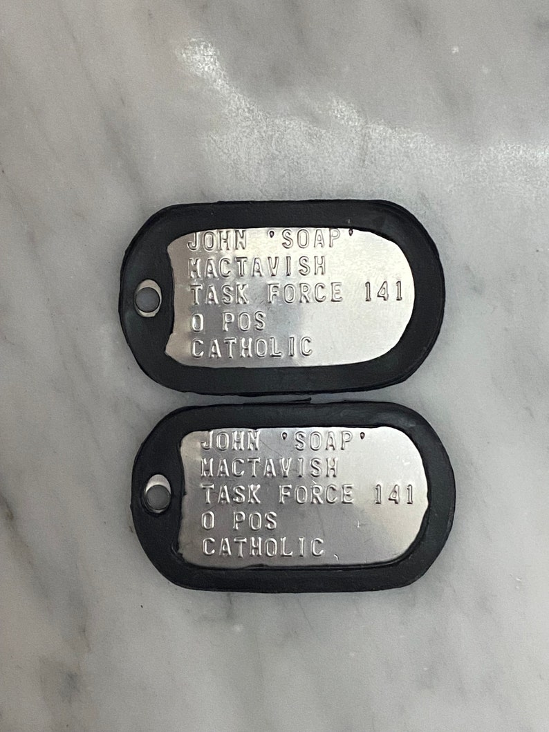 May include: Two silver dog tags with black rubber edging. The text on the tags reads "JOHN SOAP MACTAVISH TASK FORCE 141 O POS CATHOLIC".