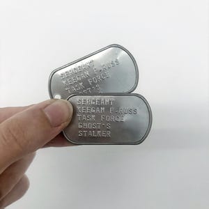 Puede incluir: Dos placas de identificación militar plateadas se muestran sobre un fondo blanco. Las placas están grabadas con el texto "SERGEANT KEEGAN P. RUSS TASK FORCE GHOST'S STALKER". Las placas tienen forma ovalada con un pequeño agujero en la parte superior.