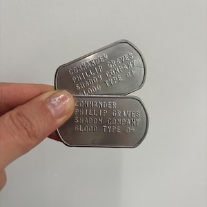 Peut inclure: Deux plaques d'identité en argent avec le texte "Commander Phillip Graves Shadow Company Blood Type 0+" dessus.