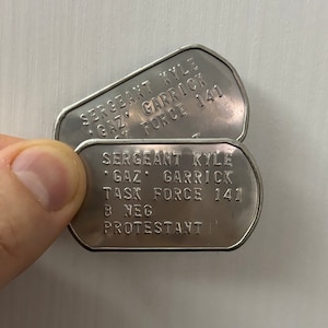 Pode incluir: Duas etiquetas de identificação militares de prata com o texto "SERGEANT KYLE GAZ GARRICK TASK FORCE 141 B NEG PROTESTANT".