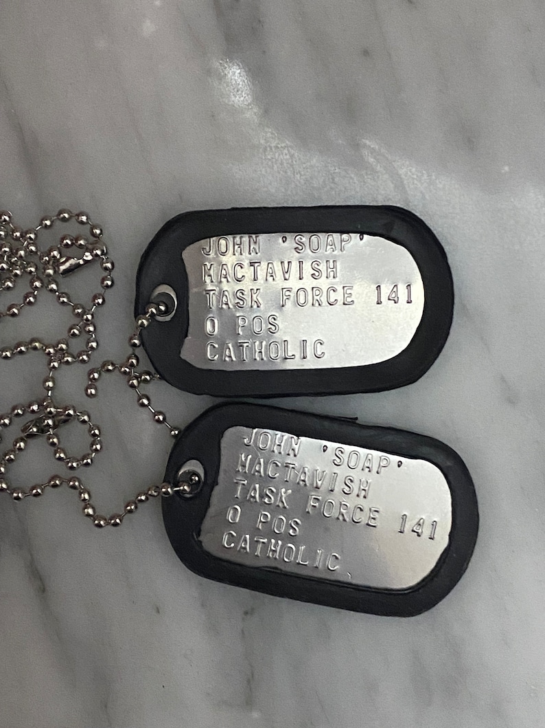 May include: Two silver dog tags with black rubber coating. The tags have the text "JOHN 'SOAP' MACTAVISH TASK FORCE 141 O POS CATHOLIC" engraved on them.