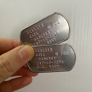 Può includere: Due targhette militari in argento con il testo "SERESIN JAKE O POS HANGHAN 247-62-7298 U.S. NAVY" su di esse.