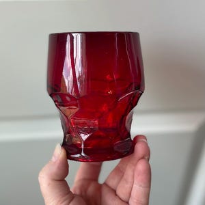 May include: A deep red glass tumbler featuring a faceted, geometric texture across its exterior surface. The vessel has a cylindrical shape that tapers slightly toward the base, with a smooth rim and a translucent, vibrant ruby-colored finish.