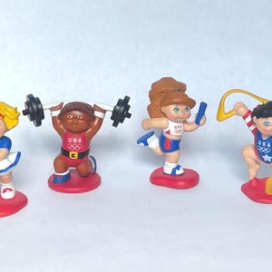 May include: Four Cabbage Patch Kids Olympic figurines standing on red bases. One holds a green tennis racket, one lifts a black barbell, one runs with a blue baton, and one performs with a yellow ribbon. Each wears a USA Olympic uniform.