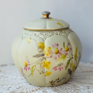 May include: A cream-colored ceramic jar with a lid, adorned with hand-painted yellow and pink floral designs. The jar has a rounded shape with a decorative rim and a gold-trimmed lid. The lid is topped with a small, blue and gold finial.