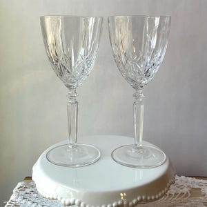 May include: Two clear crystal wine glasses with starburst and vertical cut patterns. The glasses have long stems and sit on a white, scalloped-edge cake stand. The stand is on a lace tablecloth.