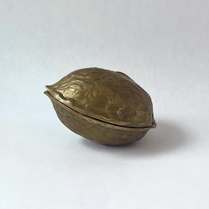 May include: A brass object shaped like a walnut with a textured, uneven surface and a hinged opening. The metal has a dull, aged gold finish with visible seams where the two halves meet, creating a realistic representation of a nut shell.