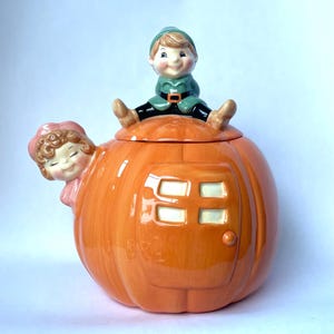 May include: A ceramic cookie jar shaped like a bright orange pumpkin house with a door and windows. A small figure with a green hat and outfit sits on the lid, while a second figure with curly hair and a pink bonnet peeks out from the side.