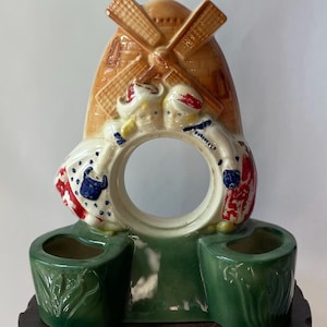 May include: A ceramic windmill-shaped planter with a brown roof and tan blades. Two figures in Dutch attire flank a circular opening. Two green planters sit below, and the piece rests on a dark wooden base. The windmill is a decorative item.