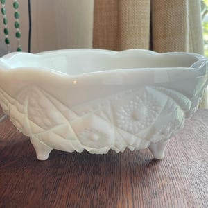 May include: A white milk glass bowl featuring a scalloped rim and a pressed geometric pattern with starburst motifs. The bowl is supported by three small feet and has a translucent quality, showcasing intricate diamond and floral-like textures.