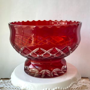 May include: A ruby red glass bowl with a scalloped rim and diamond-cut detailing. The bowl sits on a pedestal base, also in ruby red. The glass reflects light, highlighting the intricate patterns and the rich color.