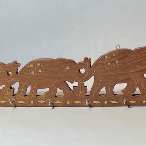 May include: A wooden key holder featuring a carved design of three elephants. The brown holder has brass accents and six metal hooks for keys. The elephants are in a row, with decorative cutouts.