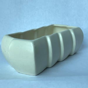May include: A rectangular off-white ceramic planter with a smooth, glossy finish. The exterior features a series of vertical, rounded ridges along the sides, creating a segmented appearance. The vessel has an open top and a hollow interior.