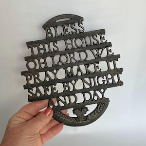 May include: A dark metal wall hanging with the words "BLESS THIS HOUSE OH LORD WE PRAY MAKE IT SAFE BY NIGHT AND DAY" in raised letters. The piece has a decorative emblem at the bottom and a handle at the top.