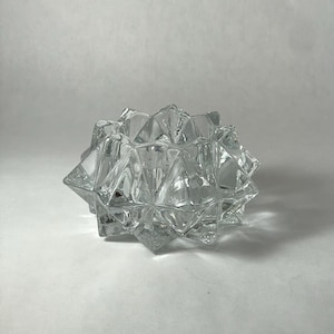 May include: A clear glass bowl featuring a multi-faceted, star-like geometric design with sharp, pointed edges and a hollow center. The transparent material reflects light, creating distinct shadows and highlights across its angular surface.