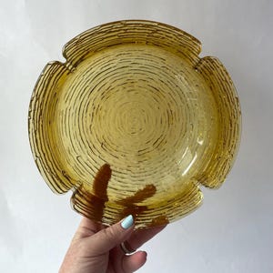 May include: A round, amber-colored glass ashtray featuring a textured, concentric ring pattern across its surface. The design includes four distinct notches along the outer rim, creating a symmetrical, circular shape with a translucent finish.