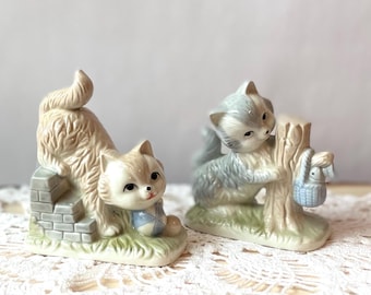 May include: Two ceramic cat figurines. One beige cat sits on a brick wall, the other gray cat climbs a tree with a blue basket. Both are on a green base, with a white background.